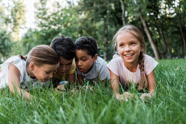 selective focus of cute multicultural children looking at green grass though magnifier 