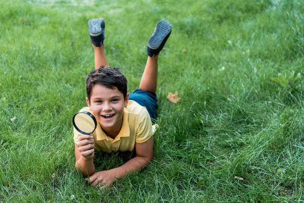cheerful boy lying on grass and holding magnifier 