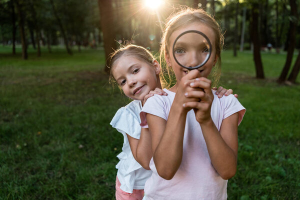happy kid holding magnifier near face while standing with friend in park 