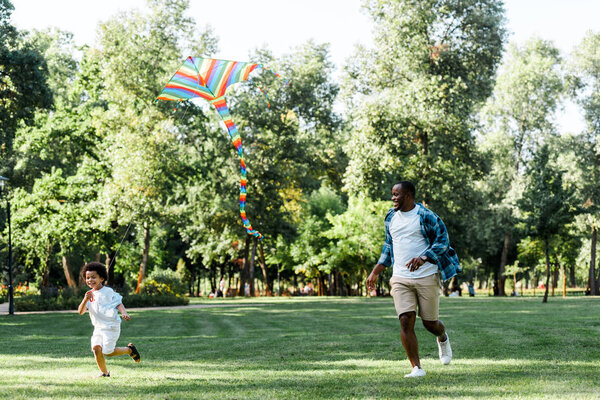 happy african american father and son running near kite in park 