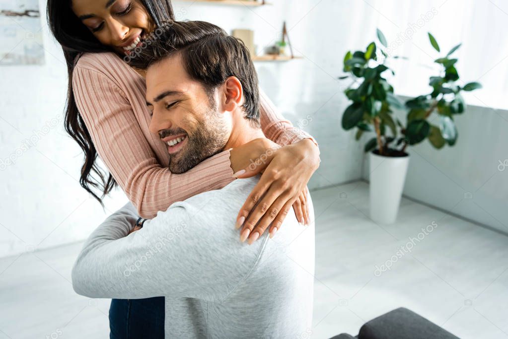 african american woman and handsome man smiling and hugging in apartment 
