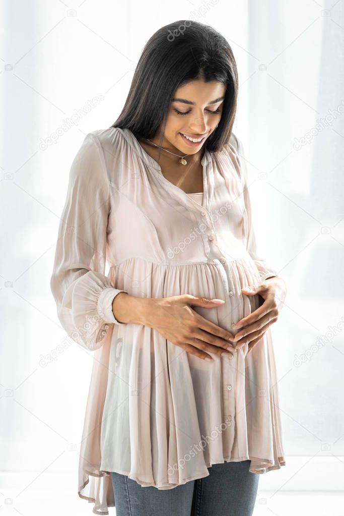 smiling pregnant african american woman hugging belly in apartment 