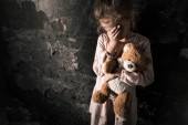 zaklatott gyerek megható arc miközben tartja teddy maci piszkos szobában, poszt apokaliptikus koncepció