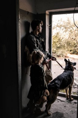 man and kid with teddy bear standing near door with german shepherd dog, post apocalyptic concept