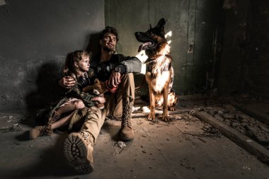 Adam ve çocuk oyuncak ayıyla yerde Alman çoban köpeğiyle oturuyor, kıyamet sonrası konsepti.