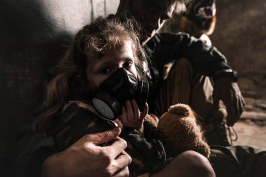 selective focus of man hugging kid in gas mask with teddy bear, post apocalyptic concept clipart