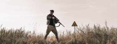 panoramic shot of handsome man holding gun near toxic symbol in field, post apocalyptic concept clipart