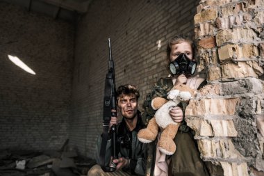 Gaz maskeli çocuk elinde oyuncak ayıyla yanında silahlı yakışıklı bir adam, kıyamet sonrası konsepti.