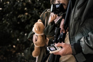 cropped view of man holding radiometer near kid in gas mask, post apocalyptic concept clipart