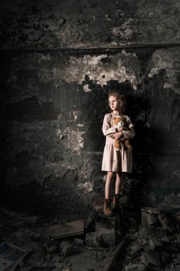 üzgün çocuk ayakta duruyor ve oyuncak ayıyı kirli odada tutuyor, kıyamet sonrası konsepti