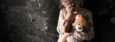 panoramic shot of upset kid touching face while holding teddy bear in dirty room, post apocalyptic concept clipart