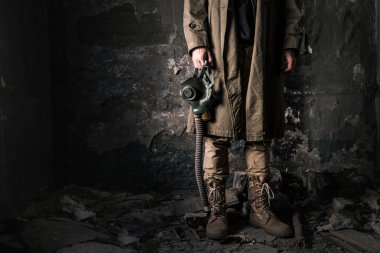 cropped view of man holding gas mask while standing near old wall in abandoned room, post apocalyptic concept clipart