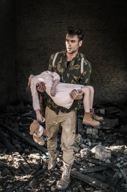 Üzgün bir adam, terk edilmiş binanın yanında oyuncak ayıyla silah tutuyor. Kıyamet sonrası konsepti.