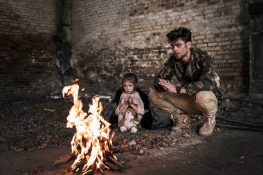 selective focus of man and child sitting near bonfire, post apocalyptic concept clipart