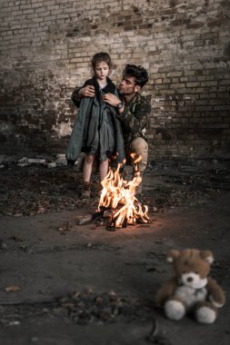 selective focus of man wearing jacket on child near bonfire, post apocalyptic concept clipart