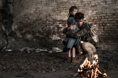 handsome man hugging child near burning bonfire, post apocalyptic concept clipart