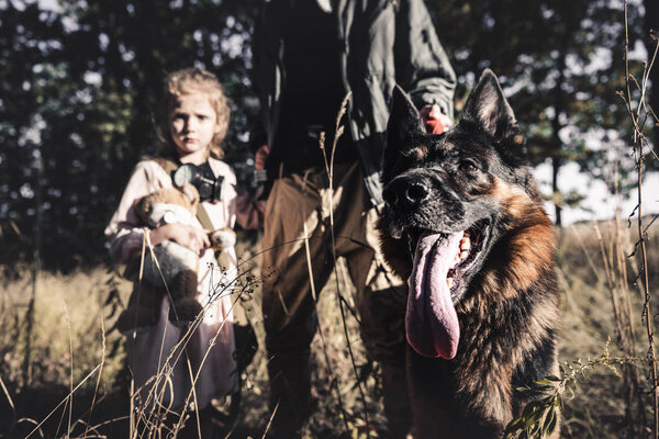 cropped view of man standing in field with cute kid and german shepherd dog, post apocalyptic concept