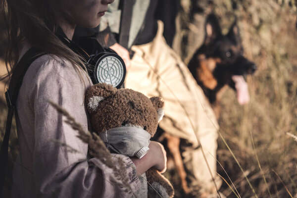 cropped view kid with gas mask holding teddy bear near man and dog, post apocalyptic concept