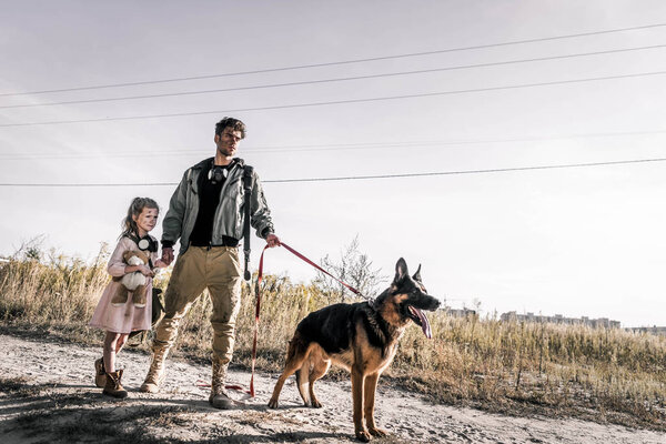 man holding hands with cute kid near german shepherd dog on road, post apocalyptic concept