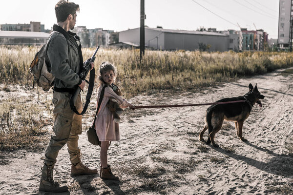 armed man holding gun near kid with teddy bear and dog, post apocalyptic concept