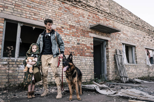 handsome man standing with kid and german shepherd dog near abandoned building, post apocalyptic concept