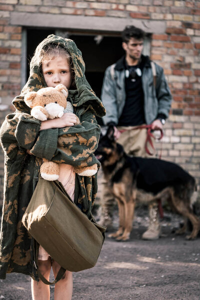 selective focus of kid holding teddy bear near man with german shepherd dog near abandoned building, post apocalyptic concept