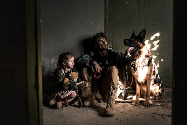 handsome man and kid with teddy bear sitting on floor with german shepherd dog, post apocalyptic concept
