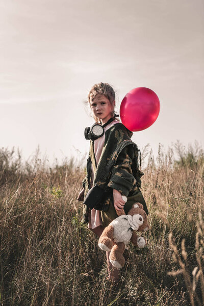 selective focus of cute kid with gas mask and teddy bear holding balloon, post apocalyptic concept