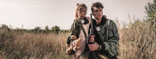 panoramic shot of handsome man holding gun near kid with soft toy in field, post apocalyptic concept