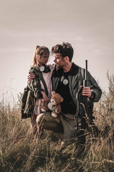 handsome man holding gun near kid with teddy bear in field, post apocalyptic concept