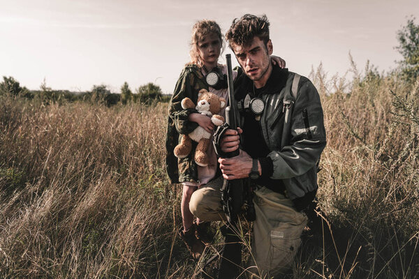 handsome man holding gun near cute kid with soft toy in field, post apocalyptic concept