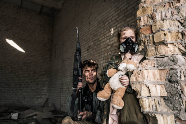 Малыш в противогазе держит плюшевого мишку рядом с красивым мужчиной с пистолетом, постапокалиптическая концепция
