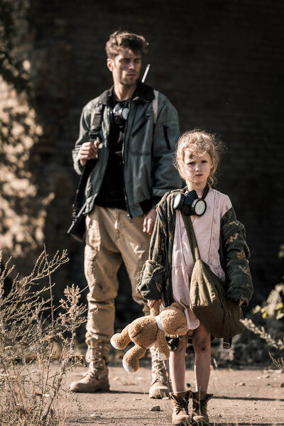 selective focus of kid with soft toy standing near armed man, post apocalyptic concept