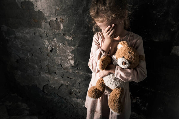 upset kid touching face while holding teddy bear in dirty room, post apocalyptic concept