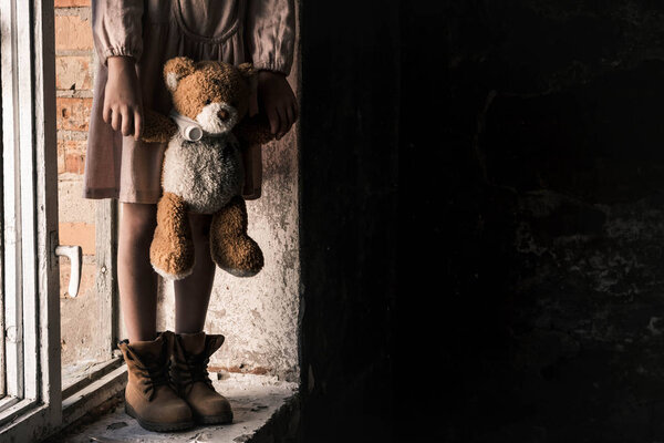 обрезанный вид ребенка, держащего грязного плюшевого мишку, стоя на подоконнике, постапокалиптическая концепция
