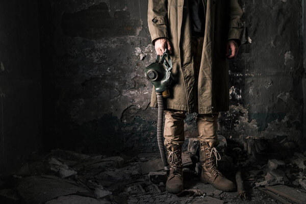 cropped view of man holding gas mask while standing near old wall in abandoned room, post apocalyptic concept