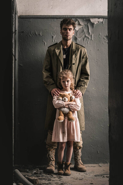 Красивый мужчина, стоящий с ребенком, держащий плюшевого мишку возле выветренной стены, постапокалиптическая концепция
