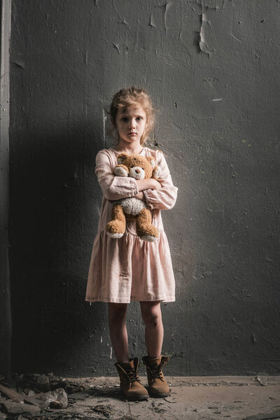 upset kid looking at camera while standing with soft toy near wall, post apocalyptic concept