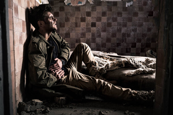 side view of man with dirty face having stomach ache while sitting on floor, post apocalyptic concept