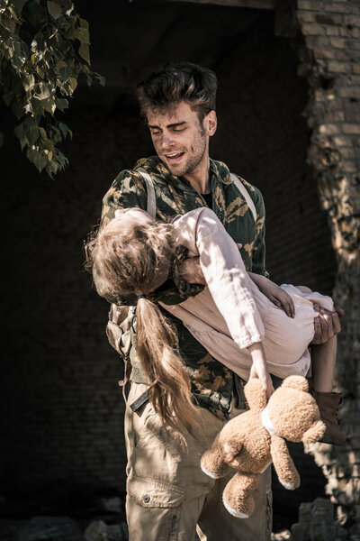 upset man holding in arms kid with teddy bear, post apocalyptic concept