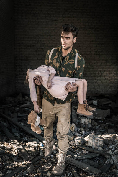 upset man holding in arms kid with teddy bear near abandoned building, post apocalyptic concept