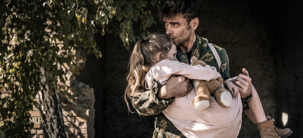 panoramic shot of man holding in arms and kissing kid with teddy bear, post apocalyptic concept