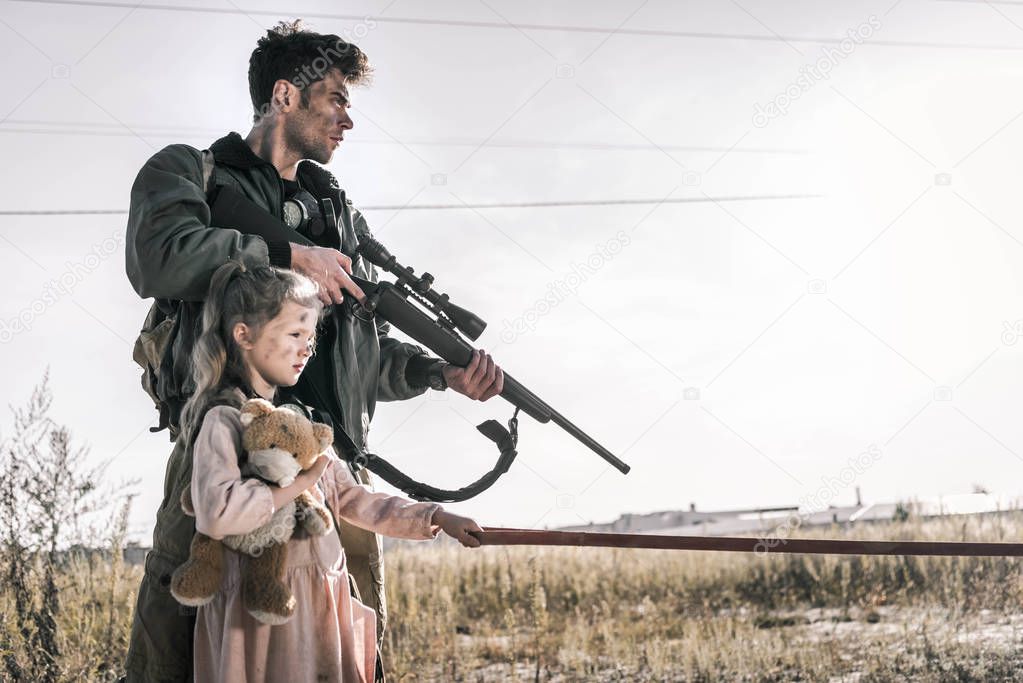 man holding gun near child with teddy bear, post apocalyptic concept