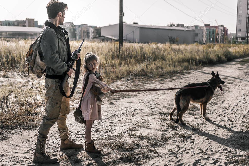 armed man holding gun near kid with teddy bear and dog, post apocalyptic concept