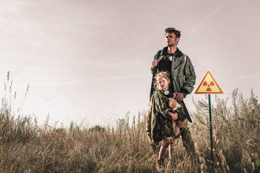 handsome man with gun near cute kid with teddy bear and toxic symbol, post apocalyptic concept