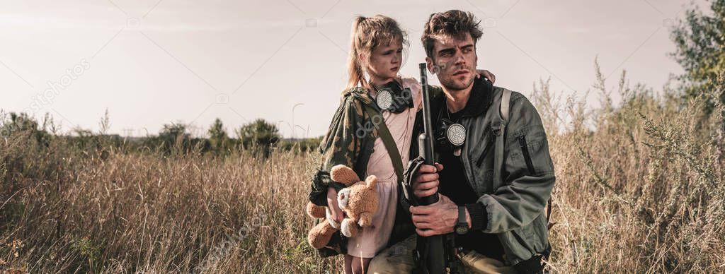 panoramic shot of handsome man holding gun near kid with soft toy in field, post apocalyptic concept