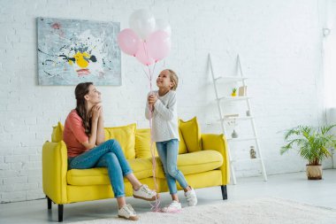 Neşeli bebek bakıcısı koltukta oturup mutlu bir çocuğun yanında pembe balonlara bakıyor. 