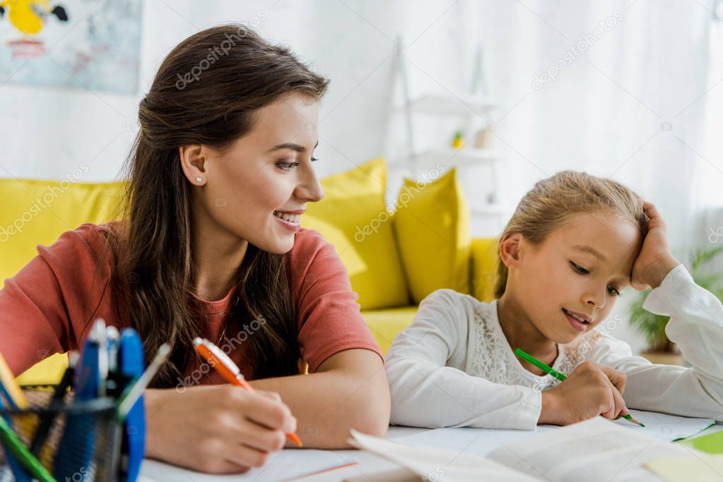 selective focus of happy babysitter looking at kid studying in living room 