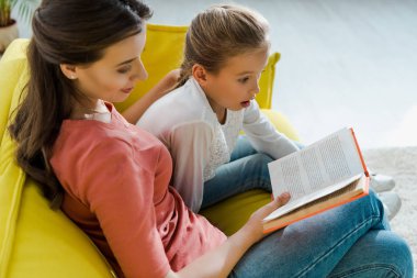 selective focus of surprised kid sitting on sofa with happy babysitter reading book  clipart