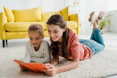 happy babysitter lying on carpet with cute kid while reading book  clipart
